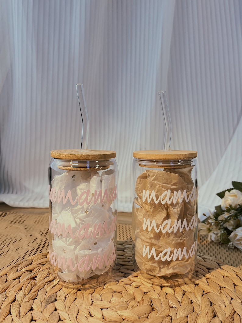 May include: Two glass tumblers with bamboo lids and pink and white straws. The tumblers have the word "mama" written on them in pink and white lettering.