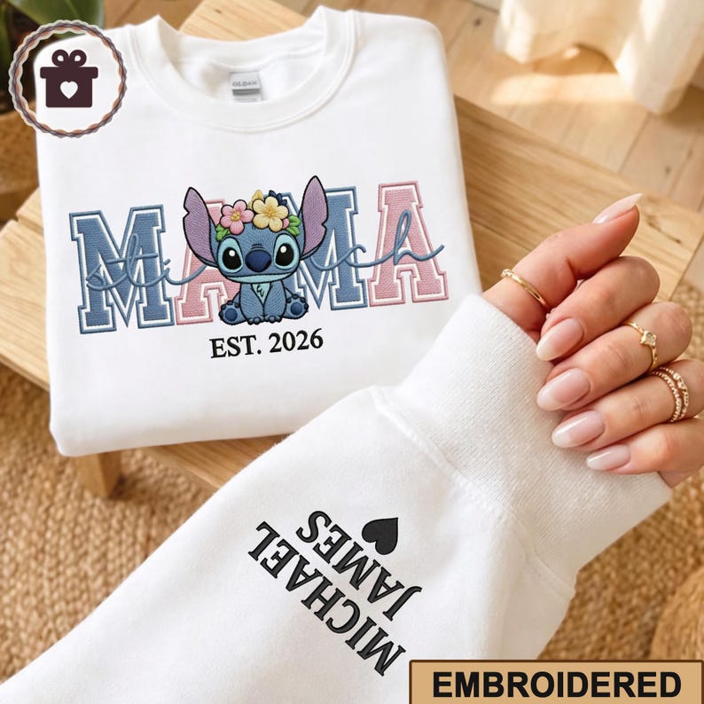 May include: White sweatshirt with embroidered "MAMA" in blue and pink, featuring a cartoon character with a floral crown. The text "EST. 2026" is below the character. The sleeve displays "MICHAEL JAMES" and a heart.