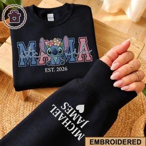 May include: Black sweatshirt with embroidered "Mama Stitch" design featuring a cartoon character with floral accents and the text "EST. 2026". The sleeve displays "MICHAEL JAMES" and a heart. The word "EMBROIDERED" is at the bottom.