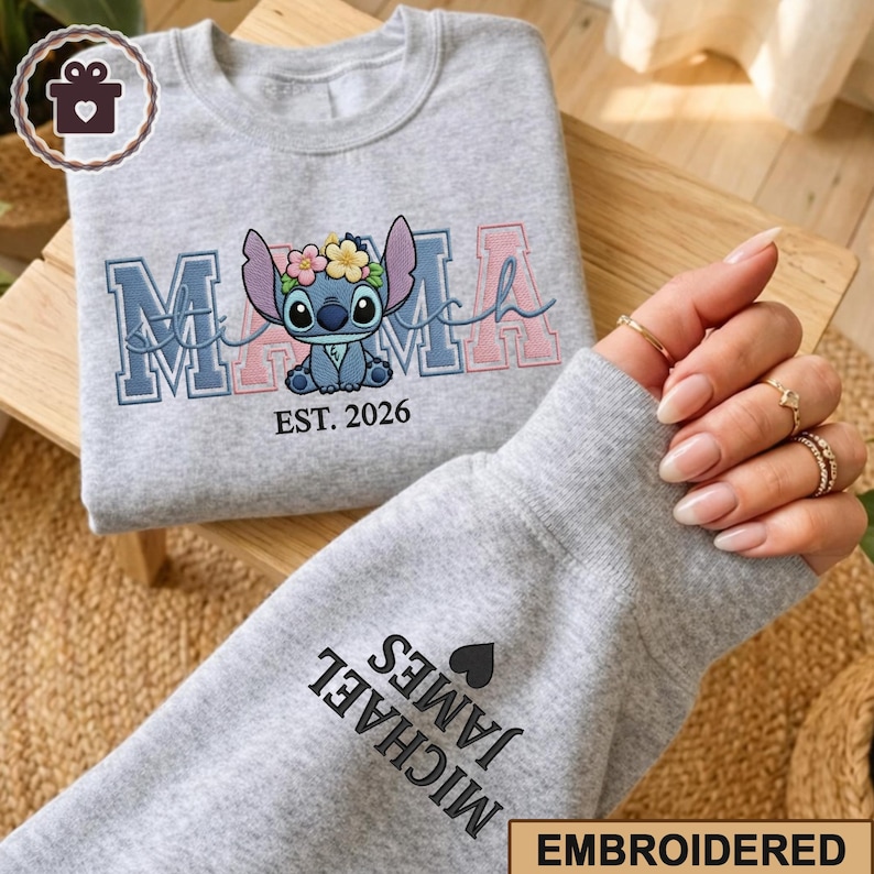 May include: A light grey sweatshirt with the word "MAMA" embroidered in blue and pink, featuring a cartoon character with a floral crown. The text "EST. 2026" is below the character. The sleeve displays the name "MICHAEL JAMES" and a heart.
