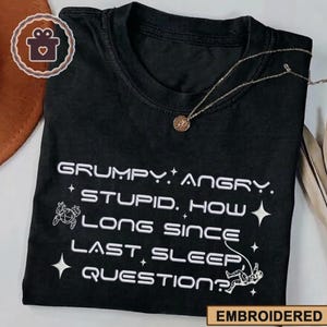 Op de afbeelding: Zwart T-shirt met witte geborduurde tekst: "GRUMPY. ANGRY. STUPID. HOW LONG SINCE LAST SLEEP, QUESTION?" Voorzien van een astronautenafbeelding en steraccenten. Het woord "EMBROIDERED" staat onderaan.