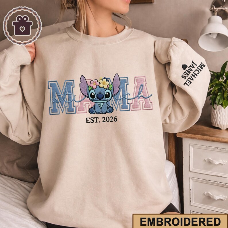 May include: Beige sweatshirt with the word "MAMA" in blue and pink embroidered letters, featuring a cartoon character with a floral crown. The text "EST. 2026" is below the character. The right sleeve has the names "MICHAEL JAMES" and a heart.