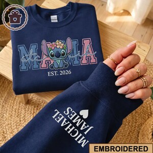 May include: Navy blue sweatshirt with embroidered "Mama Stitch" design featuring Stitch with a floral crown. The sleeve displays "MICHAEL JAMES" and a heart. "EST. 2026" is below the Stitch graphic. The word "EMBROIDERED" is at the bottom.