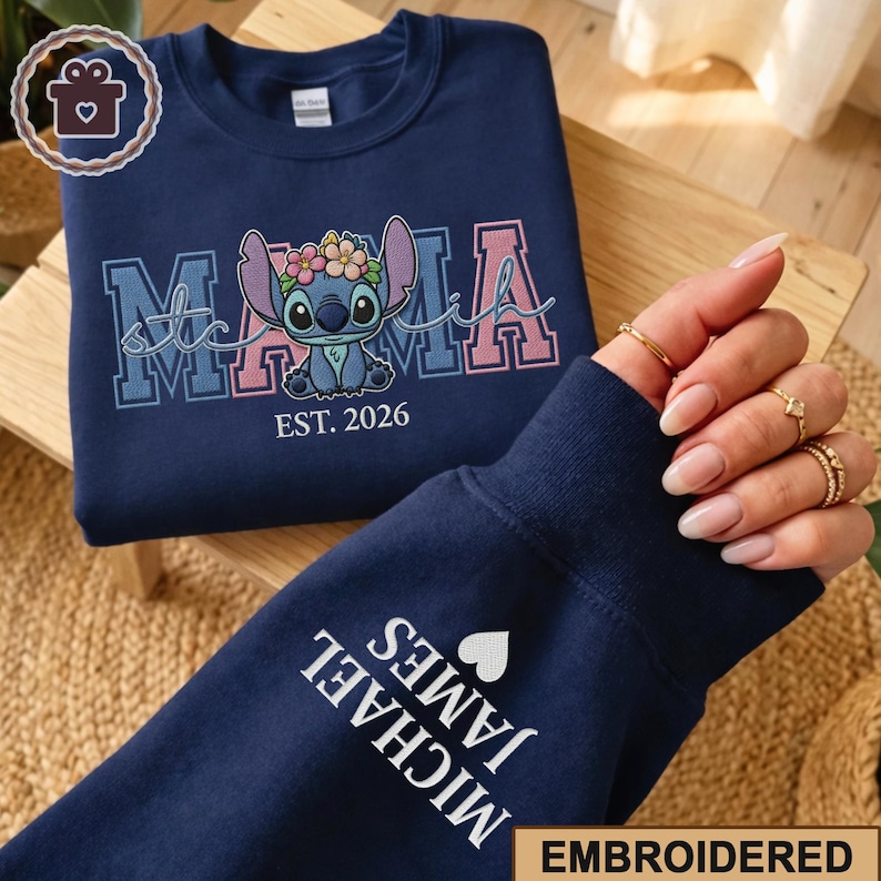 May include: Navy blue sweatshirt with embroidered "Mama Stitch" design featuring Stitch with a floral crown. The sleeve displays "MICHAEL JAMES" and a heart. "EST. 2026" is below the Stitch graphic. The word "EMBROIDERED" is at the bottom.