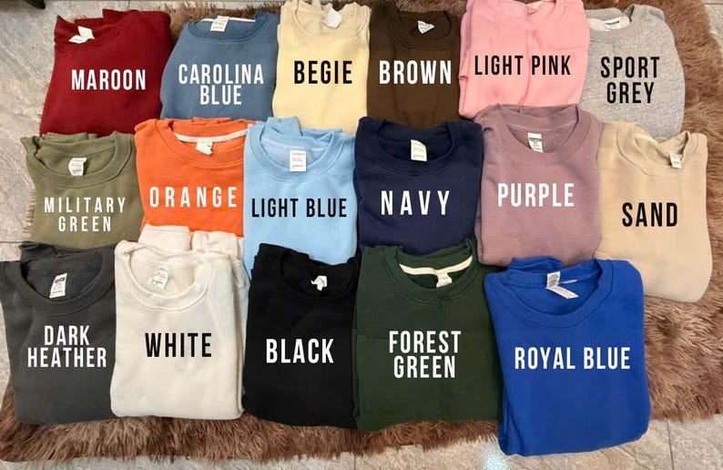May include: A collection of crewneck sweatshirts in various colors, including maroon, Carolina blue, beige, brown, light pink, sport grey, military green, orange, light blue, navy, purple, sand, dark heather, white, black, forest green, and royal blue. Each shirt has the color name printed on it.
