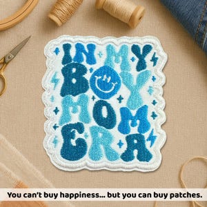 May include: Embroidered patch with the text "IN MY BOY MOM ERA" in shades of blue and white. The patch has a white scalloped border and a smiling face graphic. Additional craft supplies are visible.