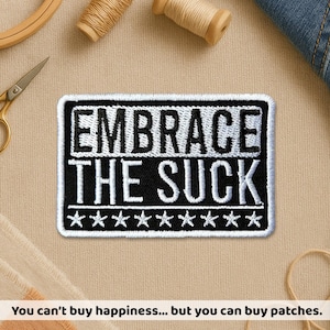 May include: Embroidered patch with the text "EMBRACE THE SUCK" in white on a black background. The patch has a white border and a row of white stars at the bottom. The patch is rectangular and is surrounded by sewing supplies.