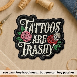 May include: Black embroidered patch with the words "Tattoos Are Trashy" in white script. The patch features a pink rose, a red rose, and a skull. The patch is surrounded by a black border and green leaves.