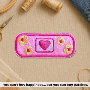 May include: A pink embroidered patch with a heart and flower design. The patch has a rectangular shape with rounded edges and a bright pink border. The center features a pink heart, surrounded by yellow flowers. The text at the bottom reads: "You can't buy happiness... but you can buy patches."