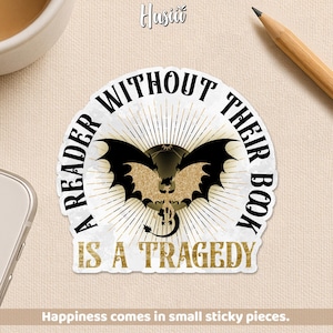 May include: A sticker with the text "A READER WITHOUT THEIR BOOK IS A TRAGEDY" in black and gold. The sticker features a bat-like creature with a skull and a dragon tail. The sticker is white with a gold sunburst design.
