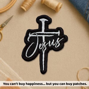 May include: Black and white embroidered patch in the shape of a cross with the word "Jesus" in cursive script. The patch is surrounded by a black border. The text at the bottom says "You can't buy happiness... but you can buy patches."