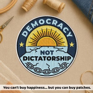 May include: A circular embroidered patch featuring a dark blue border with the text DEMOCRACY NOT DICTATORSHIP in white. The centre displays a yellow sun rising over a light blue cracked surface with a grey chain and two yellow stars.
