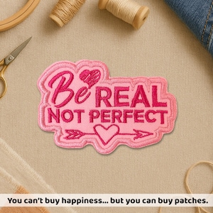 May include: A pink embroidered patch with the words "Be REAL NOT PERFECT" in a cursive and block font. The patch features a heart and arrow design. The patch is on a beige fabric surface.