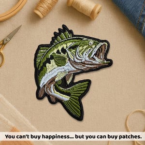 May include: Embroidered patch featuring a detailed largemouth bass in shades of green, black, white, and brown. The fish is depicted with its mouth open, showcasing intricate stitching. The patch is outlined in black. Text at the bottom reads: "You can't buy happiness... but you can buy patches."