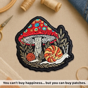 May include: Embroidered patch featuring a red mushroom with blue and yellow spots, a white stem, and two snails. The patch is surrounded by green foliage and a black border. Text at the bottom reads: "You can't buy happiness... but you can buy patches."