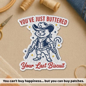 May include: Embroidered patch featuring a cartoon cat in a cowboy hat and vest, holding two pistols. The text "YOU'VE JUST BUTTERED" and "Your Last Biscuit" are above and below the cat. The patch has a white border with red and blue accents.