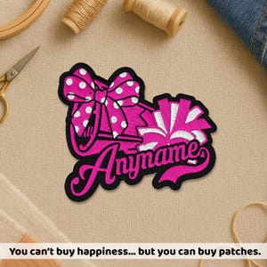 May include: An embroidered patch featuring a bright pink megaphone with a white polka-dot bow and a matching pink and white pom-pom. The word Anyname is stitched in a cursive pink font across the bottom, all outlined in a thick black border.