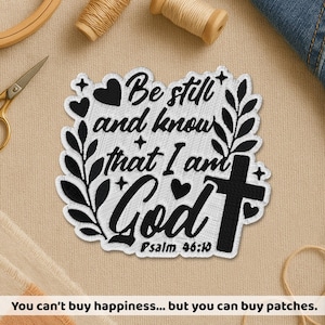 May include: Embroidered patch with the text "Be still and know that I am God" and "Psalm 46:10". The patch features a cross, hearts, and floral accents in black and white. The patch is on a beige surface.