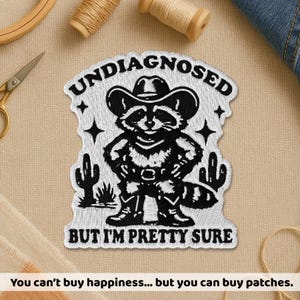 May include: Embroidered patch with a raccoon in a cowboy hat, text reads "UNDIAGNOSED BUT I'M PRETTY SURE". The patch is white with black details, including cacti and stars. Sewing supplies, including thread and scissors, surround the patch.