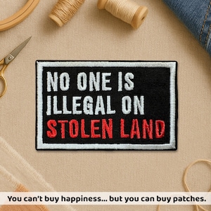 May include: A black rectangular patch with white and red text: "NO ONE IS ILLEGAL ON STOLEN LAND." The patch has a white border. The background includes sewing supplies and the text "You can't buy happiness... but you can buy patches."