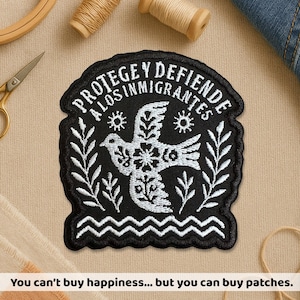 May include: Black embroidered patch with the text "PROTEGE Y DEFIENDE A LOS INMIGRANTES" and a white dove design. The patch has a wavy border and is surrounded by leaves and sunbursts. The text at the bottom reads "You can't buy happiness... but you can buy patches."