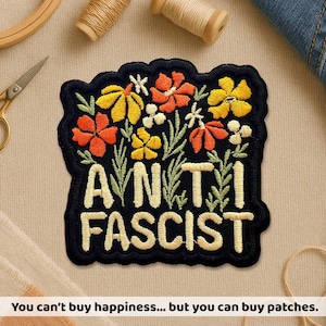 Peut inclure: Écusson brodé avec les mots "ANTI FASCISTE" en fil crème. Le patch présente un motif floral avec des fleurs orange, jaunes et blanches sur fond noir. Le patch est entouré d'une bordure noire.
