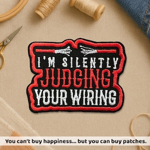May include: Embroidered patch with the text "I'M SILENTLY JUDGING YOUR WIRING" in white and red. The patch has a black background and a red border. The patch is rectangular with rounded corners. The patch is on a beige surface.