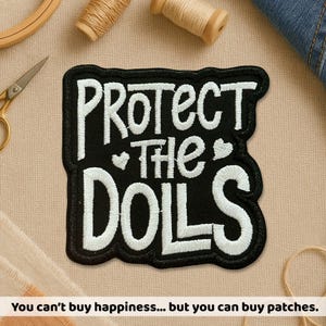 May include: Black embroidered patch with the words "PROTECT THE DOLLS" in white, with a heart symbol. The patch has a black border. The image also includes scissors, thread spools, and a wooden embroidery hoop.
