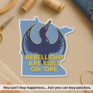 May include: Embroidered patch featuring a stylized bird design in navy blue and yellow, set against a light blue background shaped like a state. The text "REBELLIONS ARE BUILT ON 'OPE" is stitched in yellow. Sewing supplies are in the background.