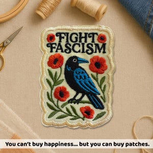 May include: Embroidered patch with the words "FIGHT FASCISM" above a black and blue bird surrounded by red poppies and green leaves. The patch has a cream-colored background and a stitched border. The text at the bottom says "You can't buy happiness... but you can buy patches."