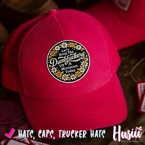 Op de afbeelding: Een felroze truckerpet met een mesh achterkant en een ronde patch. De patch bevat de tekst "Let's Keep The Dumbfuckery To A Minimum Today" omringd door een bloemenontwerp. De afbeelding bevat ook de tekst "HATS, CAPS, TRUCKER HATS".