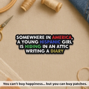 May include: A black patch with white text that reads "SOMEWHERE IN AMERICA, A YOUNG HISPANIC GIRL IS HIDING IN AN ATTIC WRITING A DIARY." The patch is surrounded by sewing supplies.
