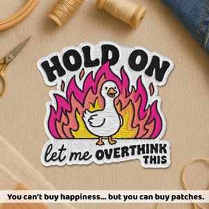 May include: Embroidered patch featuring a white goose in front of pink and orange flames. The patch reads "HOLD ON, let me OVERTHINK THIS" in black lettering. The patch is surrounded by sewing supplies.