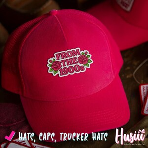May include: A vibrant pink trucker hat with a mesh back and a curved brim. The hat features a patch with the text "FROM THE 1900s" surrounded by roses and leaves. The image also includes the text "HATS, CAPS, TRUCKER HATS".