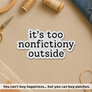 May include: Embroidered patch with the phrase "it's too nonfictiony outside" in black text on a white background. The patch is surrounded by sewing supplies, including scissors, thread spools, and a wooden embroidery hoop. The text "You can't buy happiness... but you can buy patches" is at the bottom.