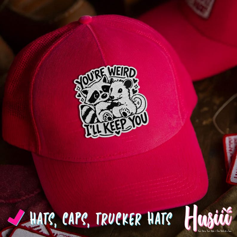 May include: A vibrant pink trucker hat with a mesh back. The front of the cap displays a patch with a raccoon and a mouse embracing, and the text "YOU'RE WEIRD I'LL KEEP YOU". The hat is part of a collection of hats and caps.