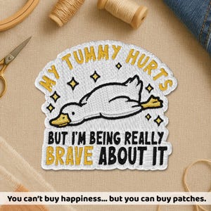 May include: Embroidered patch with the text "MY TUMMY HURTS" and "BUT I'M BEING REALLY BRAVE ABOUT IT" in yellow and black. The patch features a cartoon duck with a yellow beak and feet. The patch is white with a yellow border.