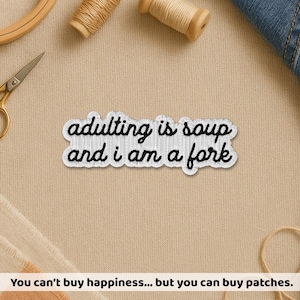 May include: Embroidered patch with the text "adulting is soup and i am a fork" in black script on a white background. The patch is surrounded by a black border. The patch is on a beige fabric surface.