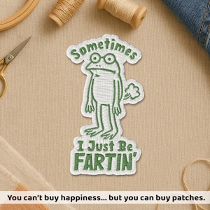 May include: Embroidered patch with a green frog design and the words "Sometimes I Just Be Fartin'". The patch has a white border and is on a tan background. Sewing supplies are also visible in the image.