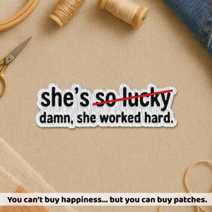 May include: Embroidered patch with the text "she's so lucky" crossed out and replaced with "damn, she worked hard." The patch is white with black lettering and a red line. The patch is on a beige surface.