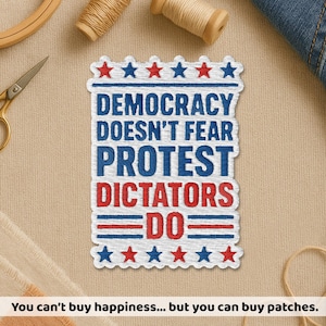 May include: Embroidered patch with the text "DEMOCRACY DOESN'T FEAR PROTEST DICTATORS DO" in blue and red. The patch has a white border with red and blue stars. The patch is surrounded by sewing supplies.