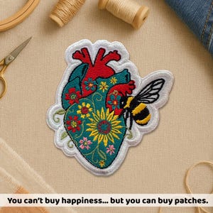 May include: Embroidered patch in the shape of a human heart, featuring a teal background with floral designs and a bee. The heart has red and teal sections, with a white border. Text at the bottom reads: "You can't buy happiness... but you can buy patches."