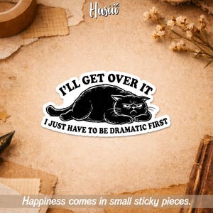 May include: A white sticker with the text "I'LL GET OVER IT I JUST HAVE TO BE DRAMATIC FIRST" and a black cat illustration. The sticker is on a tan background with other decorative elements.