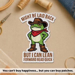 May include: Embroidered patch featuring a green frog wearing a cowboy hat, red bandana, and boots. The patch reads "MIGHT BE LAID BACK BUT I CAN LEAN FORWARD READ QUICK". The patch is white with brown and green accents.
