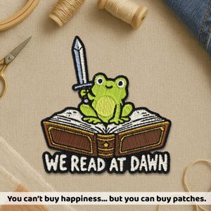 May include: An embroidered patch featuring a light green frog sitting on an open brown book, holding a silver sword. The text "WE READ AT DAWN" is embroidered in white letters at the bottom of the patch, outlined in black thread.