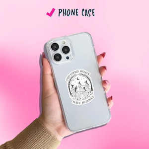 May include: Clear phone case with a black and white graphic of three witches dancing in a circle, with the text "GOD FORBID WOMEN HAVE HOBBIES". The phone case is held in a hand against a pink background.