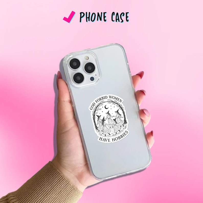 May include: Clear phone case with a black and white graphic of three witches dancing in a circle, with the text "GOD FORBID WOMEN HAVE HOBBIES". The phone case is held in a hand against a pink background.