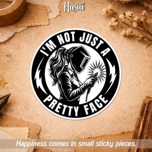 May include: A round black and white sticker with the text "I'M NOT JUST A PRETTY FACE". The sticker depicts a female welder in a welding mask, holding a torch and creating sparks. The design is bold and graphic, suitable for various applications.