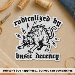 May include: Embroidered patch with the text "radicalized by basic decency" and a black and white illustration of an angry cat. The patch is white with black text and a black cat design. The image also includes the text "You can't buy happiness... but you can buy patches."