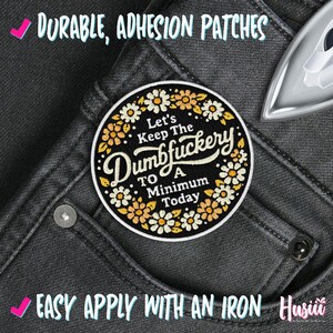 Op de afbeelding: Een ronde, geborduurde patch met de tekst "Let's Keep The Dumbfuckery To A Minimum Today" omringd door bloemen. De patch bevindt zich op een grijze jeans. De woorden "Durable, Adhesion Patches" en "Easy Apply With An Iron" zijn ook zichtbaar.